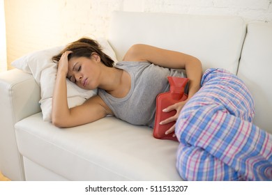 Young Beautiful Hispanic Woman In Painful Expression Holding Hot Water Bottle Against Belly Suffering Menstrual Period Pain Lying Sad On Home Couch Having Tummy Cramp In Female Health Concept