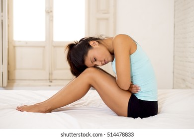 young beautiful hispanic woman in painful expression holding her belly suffering menstrual period pain lying sad on home bed having tummy cramp in female health concept