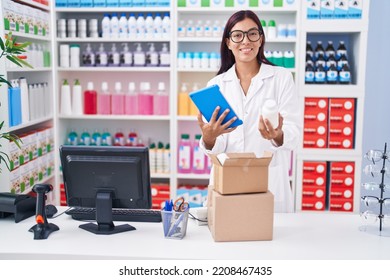 Young Beautiful Hispanic Woman Pharmacist Using Touchpad Holding Pills Bottle At Pharmacy