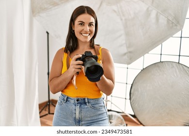 Young beautiful hispanic woman photographer holding professional camera photo studio