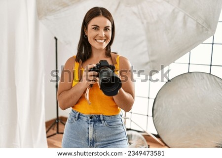 Young beautiful hispanic woman photographer holding professional camera photo studio