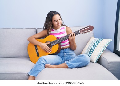 Young Beautiful Hispanic Woman Playing Classical Guitar Sitting On Sofa At Home