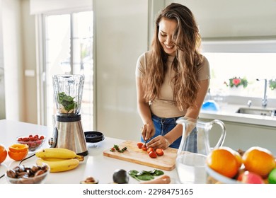 Young beautiful hispanic woman preparing vegetable smoothie with blender cutting strawberries at the kitchen - Powered by Shutterstock