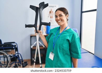 Joven y hermosa mujer hispana pisioterapeuta sonriente confiada sosteniendo muletas en la clínica de rehabilitación