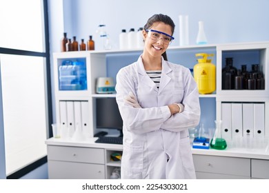 Joven y hermosa científica hispana sonriendo confiada de pie con los brazos cruzados gesto en el laboratorio