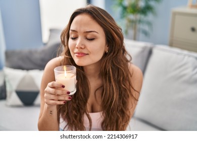 Young beautiful hispanic woman sitting on sofa smelling aromatic candle at home