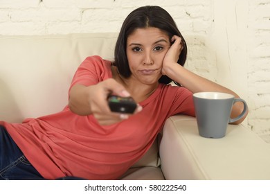 young beautiful hispanic woman sitting at home sofa couch in living room watching television looking tired and bored disappointed holding remote control switching channels