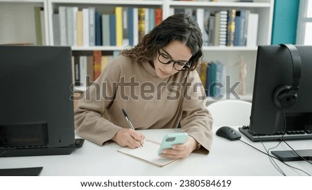 Young beautiful hispanic woman student using smartphone taking notes at library university