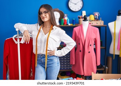 Young Beautiful Hispanic Woman Tailor Smiling Confident Leaning On Clothes Rack At Clothing Factory