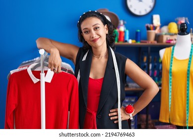 Young Beautiful Hispanic Woman Tailor Smiling Confident Leaning On Clothes Rack At Clothing Factory