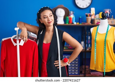 Young Beautiful Hispanic Woman Tailor Smiling Confident Leaning On Clothes Rack At Clothing Factory