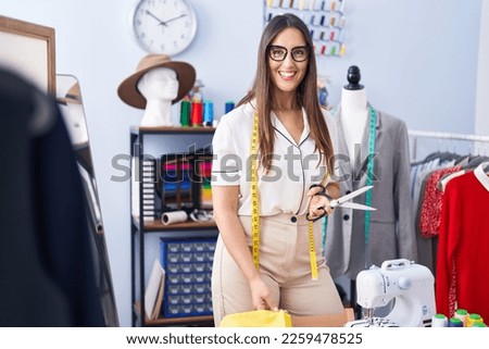 Young beautiful hispanic woman tailor smiling confident cutting cloth at clothing factory