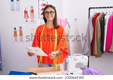Young beautiful hispanic woman tailor smiling confident looking clothing design at tailor shop