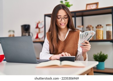 Young beautiful hispanic woman using laptop counting dollars at home