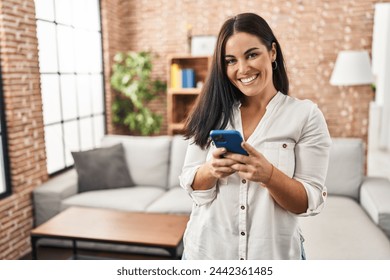 Young beautiful hispanic woman using smartphone standing at home