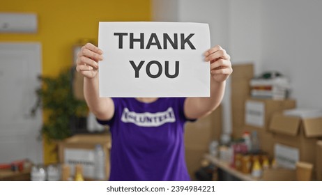 Young beautiful hispanic woman volunteer holding thank you message over face at charity center
