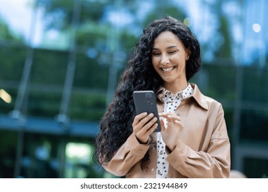 Young beautiful hispanic woman walking in the city, business woman holding phone in hands using smartphone app, woman smiling contentedly and happy outside office building with curly hair - Powered by Shutterstock