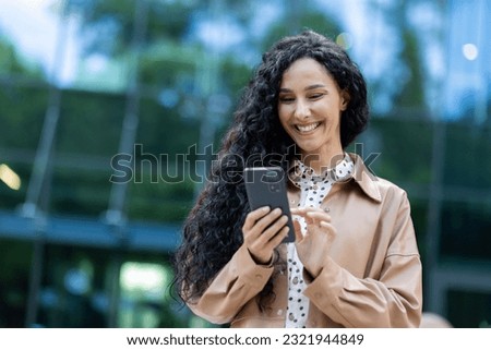 Young beautiful hispanic woman walking in the city, business woman holding phone in hands using smartphone app, woman smiling contentedly and happy outside office building with curly hair