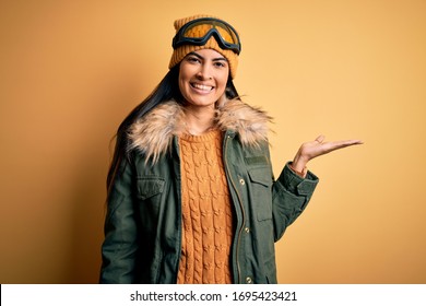 Young beautiful hispanic woman wearing ski glasses and coat for winter weather smiling cheerful presenting and pointing with palm of hand looking at the camera.