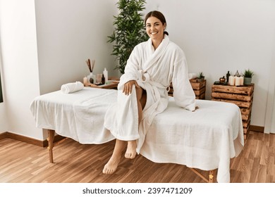 Young beautiful hispanic woman wearing bathrobe sitting on massage table at beauty salon