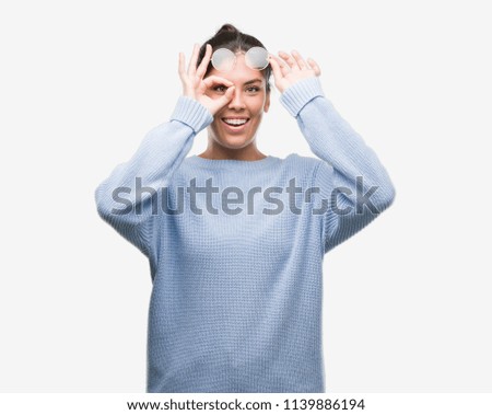 Young beautiful hispanic woman wearing a bun with happy face smiling doing ok sign with hand on eye looking through fingers