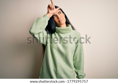 Young beautiful hispanic woman wearing green winter sweater over isolated background making fun of people with fingers on forehead doing loser gesture mocking and insulting.