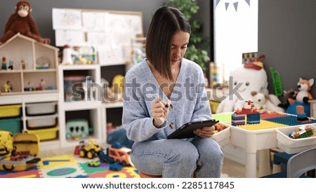 Young beautiful hispanic woman working as a teacher talking on smartphone using touchpad at kindergarten