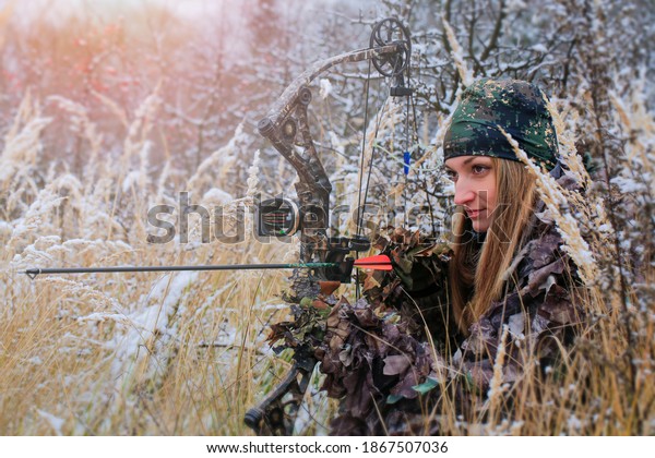 A young beautiful Hunter woman in camouflage in\
snowy forest with hunting bow in her hand. Winter lanscape. Copy\
space for text.