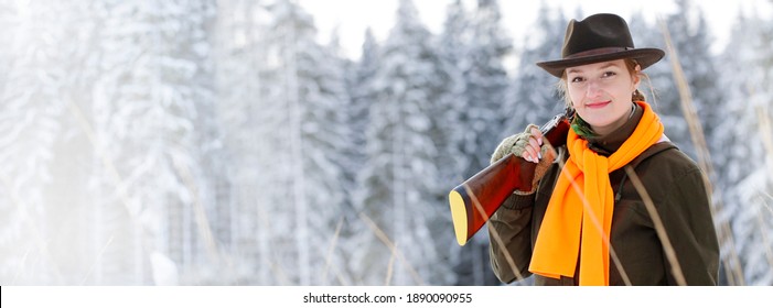 A young beautiful hunter woman on hunt in forest with rifle on the shoulder. Winter season. In the background are snowy trees. Banner photo.
