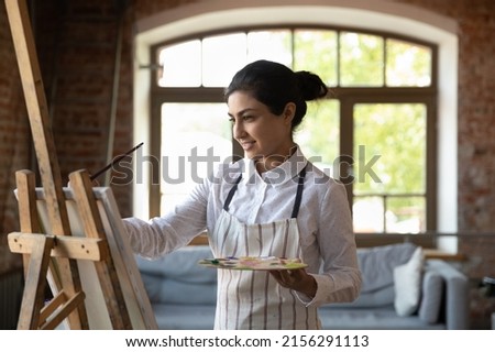 Young beautiful Indian woman painter creates pictures, drawing with paints on canvas in modern workshop. Professional occupation, creative hobby, activity for soul, vocation, develop skills concept