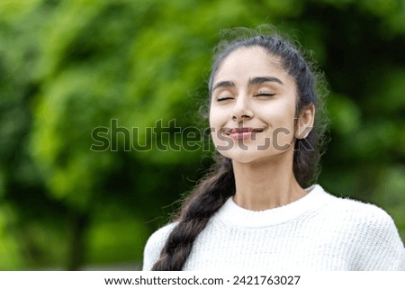 A young beautiful Indian woman is standing outside in a park in a white sweater and with her eyes closed is taking deep breaths and enjoying herself. Close-up photo.