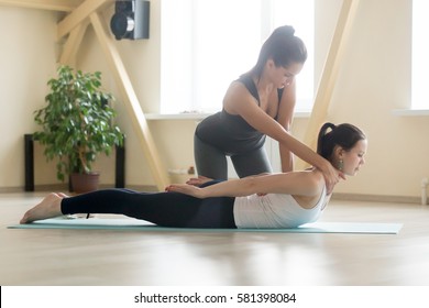 Young beautiful lady beginning yoga practice with private teacher at home class, working out with professional female yogi instructor. Trainer helps student to do Salabhasana exercise, Locust pose 