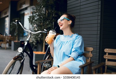 Young beautiful lady drinking fresh smoothies on a sunny summer day during her ride. Riding a bicycle in the city. Bicycle tourism. Refreshing fruit drink. Active rest. Hipster on a bike.