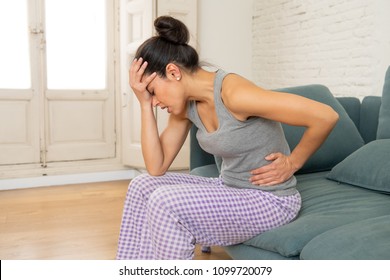 Young Beautiful Latin Hispanic Woman In Painful Expression Suffering Head Ache, Stomach Cramps And Menstrual Period Pain Sitting Sad In The Sofa At Home. Woman Female Health Concept