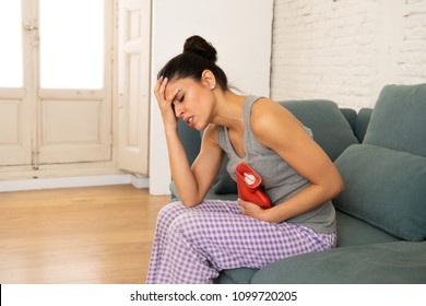 Young Beautiful Latin Hispanic Woman In Painful Expression Suffering Stomach Cramps And Menstrual Period Pain Holding A Hot Water Bottle Against Belly Sitting In The Sofa. Woman Health Concept