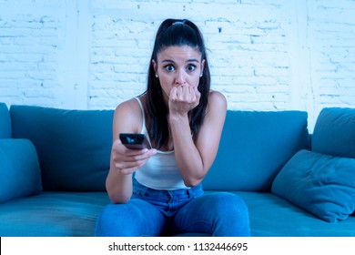 young beautiful latin woman sitting at home sofa couch in living room watching television scary horror movie or horrible news scared and excited covering her eyes holding remote control.