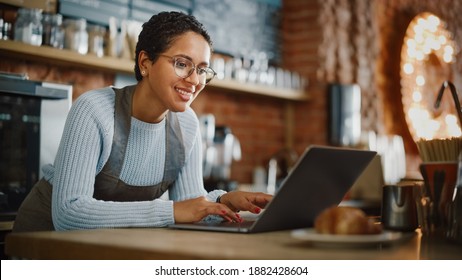 Young and Beautiful Latina Coffee Shop Owner is Working on Laptop Computer and Checking Inventory in a Cozy Cafe. Successful Restaurant Manager Browsing Internet and Accepting Online Take Away Orders.