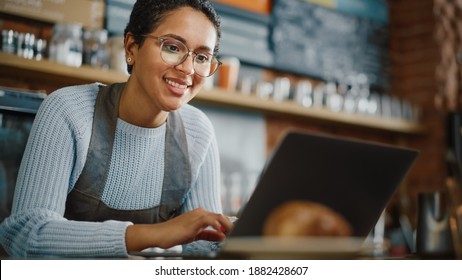 Young And Beautiful Latina Coffee Shop Owner Is Working On Laptop Computer And Checking Inventory In A Cozy Cafe. Happy Restaurant Manager Or Employee Browsing Internet And Chatting With Friends.