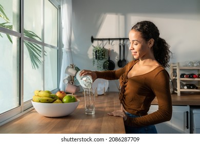 Young beautiful Latino woman pouring clean water into glass in kitchen. Attractive active thirsty girl drink or take a sips of mineral natural in cup for health care and wellbeing in kitchen in house. - Shutterstock ID 2086287709