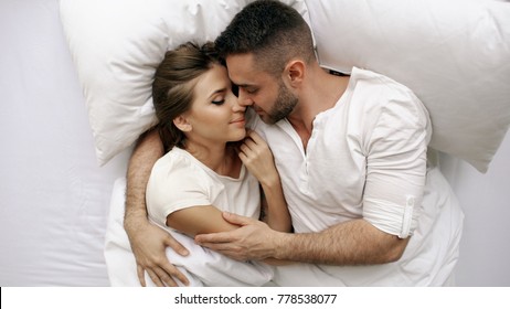 Young beautiful and loving couple kiss and hug into bed while waking up in the morning. Top view of attractive man kissing and talking his smiling wife