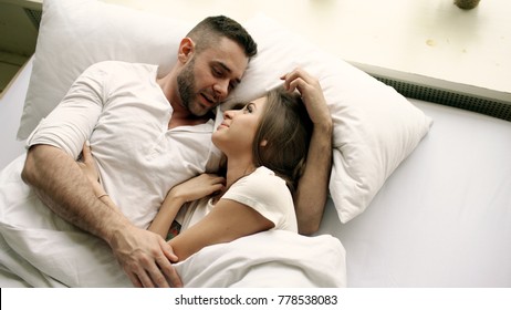 Young beautiful and loving couple talk and hug into bed while waking up in the morning. Top view of attractive man chatting his smiling wife