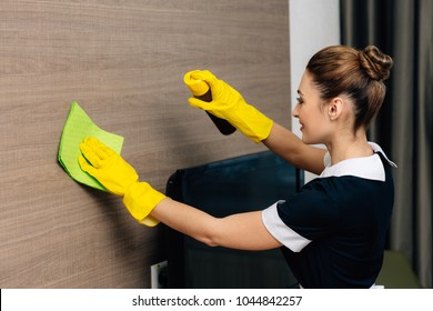 Young Beautiful Maid In Uniform Wiping Wood Wall With Rag And Aerosol Furniture Cleaner