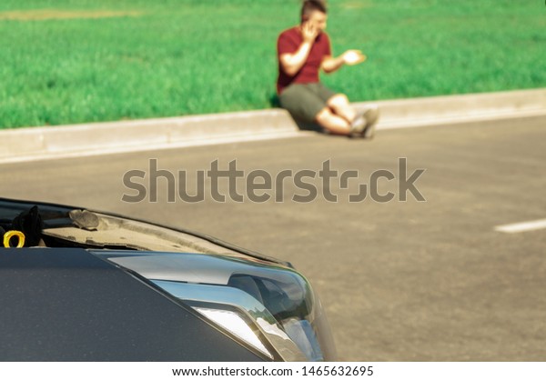 Young and beautiful man near a broken car with an\
open hood, talking on the phone. Problems with the machine, does\
not start, does not work