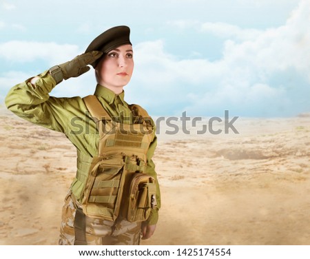 Young beautiful military soldier woman in protective armor tactical vest, camouflage pants, military beret and gloves, standing and saluting on desert background.