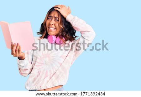 Young beautiful mixed race woman reading a book wearing headphones stressed and frustrated with hand on head, surprised and angry face 