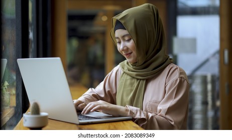 Young Beautiful Modern Asian Muslim Women Sitting By The Window Enjoying A Relaxing Moment Working On A Laptop In The Coffee Shop On A Bright Sunny Day