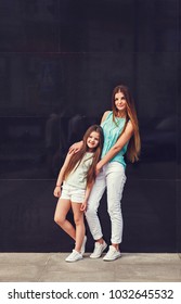 Young Beautiful Mom With Her Daughter Wearing T-shirt And Jeans Posing Against Concrete Wall, Minimalist Street Fashion Style, Clothing For Parent And Child.