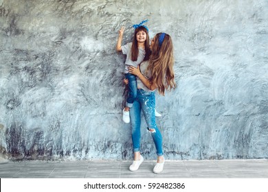 Young Beautiful Mom With Her Daughter Wearing Blank Gray T-shirt And Jeans Posing Against Rough Concrete Wall, Minimalist Street Fashion Style, Family Same Look, Clothing For Parent And Child.