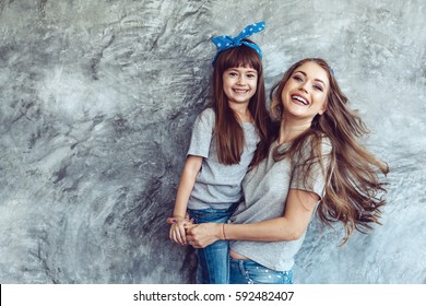 Young Beautiful Mom With Her Daughter Wearing Blank Gray T-shirt And Jeans Posing Against Rough Concrete Wall, Minimalist Street Fashion Style, Family Same Look, Clothing For Parent And Child.