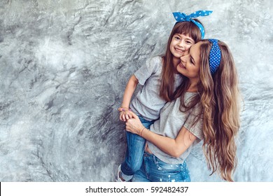 Young Beautiful Mom With Her Daughter Wearing Blank Gray T-shirt And Jeans Posing Against Rough Concrete Wall, Minimalist Street Fashion Style, Family Same Look, Clothing For Parent And Child.